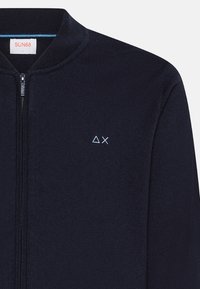 Cardigan zip-up blu navy realizzato in tessuto a maglia, con colletto alto e piccolo logo azzurro a sinistra sul petto. Texture liscia, design aderente.