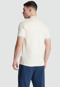 Cremfarbener Poloshirt mit kurzen Ärmeln, ausgestattet mit einem gerippten Kragen und dunkelblauen Streifen. Glatter Stoff mit klassischem Schnitt, kombiniert mit dunkelblauen Hosen.