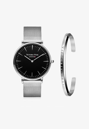 Reloj con esfera negra y correa de malla plateada, que presenta marcadores de hora y agujas delgadas. Acompañado de una pulsera de puño plateada grabada con el nombre de la marca.