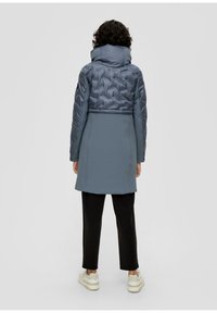 Helles blauer gepolsterter Mantel mit hohem Kragen, gestepptem Obermaterial und glattem Stoff darunter. Kombiniert mit schwarzen schmal zulaufenden Hosen und weißen Turnschuhen.