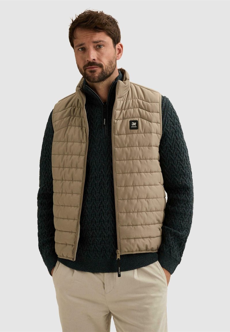 Homme aux cheveux bruns et barbe portant une veste matelassée beige sur un pull à torsades vert foncé et un pantalon de couleur claire sur fond uni.