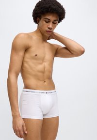Λευκά βαμβακερά ανδρικά εσώρουχα boxer με μαύρη μέση που φέρει το λογότυπο "TOMMY HILFIGER", με εφαρμοστή γλώσσα και λεία υφή. Το μοντέλο ποζάρει πλάγια.