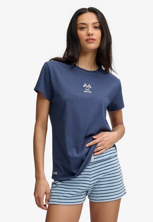Kvinde iført en mørkeblå t-shirt med en lille hvid logo og lyseblå shorts med sorte vandrette striber, stående mod en hvid baggrund.