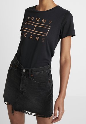Frau trägt ein schwarzes Tommy Jeans T-Shirt mit kupferfarbenem Logo und einen schwarzen, abgenutzten Jeansrock.