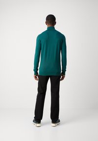 Pull turquoise à manches longues avec un col montant, coupe ajustée et poignets côtelés, associé à un pantalon noir et des baskets multicolores.