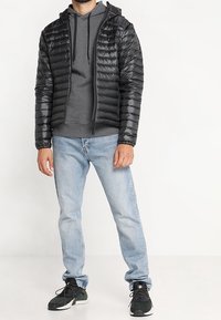 Chaleco acolchado negro con capucha, usado sobre una sudadera gris. Jeans azul claro y zapatillas deportivas negras completan el look. Textura suave y brillante.