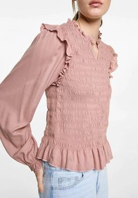 Rosa, texturerad blus med flikiga axlar och en böljande fåll. Har en V-ringning och långa ärmar av lättviktsmaterial.