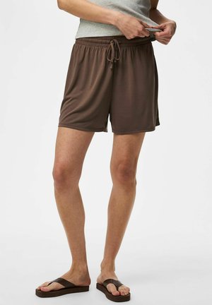 Person iført brune shorts med snøre, grå ærmeløs top og brune flip-flops, står mod en ensfarvet baggrund.