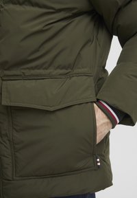 Grön puffjacka med en framficka, med stickade manschetter med röda, vita och blå ränder, tillverkad av ett slätt, vattenavvisande material.