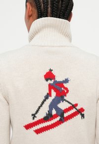 Pull à col roulé en tricot beige arborant un motif de skieur coloré en rouge, bleu et noir au dos sur fond neutre.