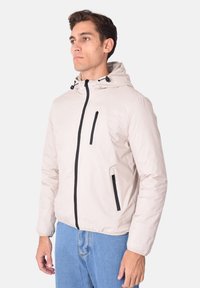 Giacca leggera beige con cappuccio, dotata di tasche con cerniera nera e dettagli in nero, texture liscia e design aderente.