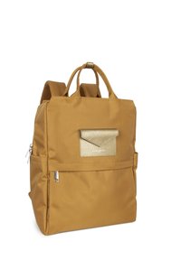 Sac à dos de taille moyenne en tissu jaune moutarde. Il dispose d'une poche à rabat en or texturé, de fermetures éclair et de bretelles rembourrées.