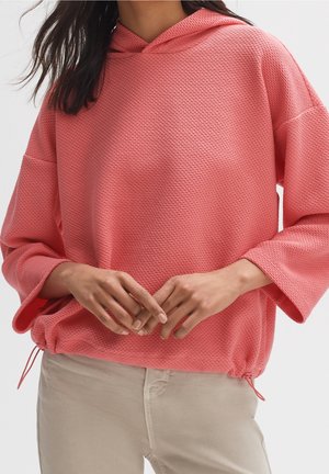 Femme portant un sweat à capuche texturé rose corail à manches longues avec cordon de serrage à l'ourlet et un pantalon beige, mains posées légèrement jointes devant.