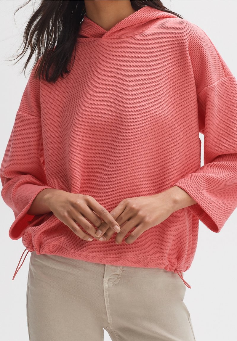 Femme portant un sweat à capuche texturé rose corail à manches longues avec cordon de serrage à l'ourlet et un pantalon beige, mains posées légèrement jointes devant.