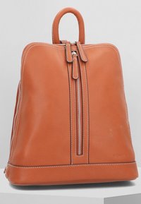Mochila de cuero marrón con dos cremalleras verticales, costuras blancas a lo largo de los bordes, un asa en la parte superior y una base plana apoyada sobre una superficie blanca.