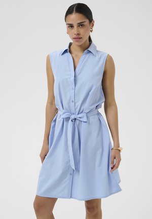 Nő világoskék, ujjatlan, gombos, kötött övű ruhában, arany karika fülbevalóval és karkötővel, egyszínű háttér előtt áll.