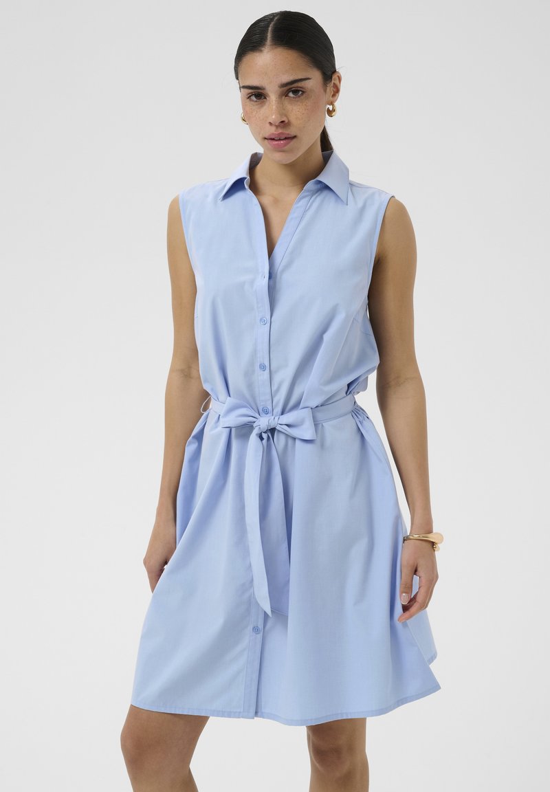 Femme portant une robe sans manches bleu clair boutonnée avec ceinture nouée, avec des boucles d'oreilles créoles dorées et un bracelet, debout devant un fond uni.