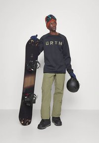 Mann in Winterkleidung, der ein Snowboard mit einer Hand senkrecht hält und einen schwarzen Helm in der anderen, während er eine Skibrille und Handschuhe trägt.