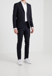 Marineblauer Anzug mit einreihiger Jacke und schmal geschnittenen Hosen, kombiniert mit einem weißen Hemd und weißen Sneakers. Glatte Stoffstruktur.