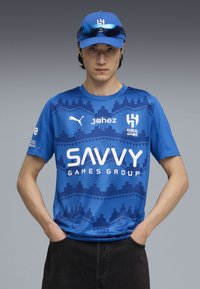 Blå sportsjersey med geometriske mønstre, prydet med hvid tekst, der er parret med en matchende blå kasket og solbriller. Mørke bukser fuldender outfittet.
