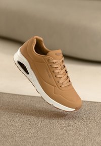 Tan syntetiska sneakers med perforerade detaljer, vit gummisula och platta snören. Har en synlig luftkuddesystem på sidan.
