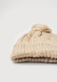 Beige kudumine villane beanie, millel on volditud äär ja pom-pom ülaosas, lamab tasase pinna peal valge taustaga.