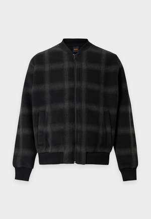 Jeune homme portant une veste bomber à carreaux noir et vert foncé, un pantalon marron foncé, une chemise claire et des chaussures en cuir noires, debout les mains dans les poches.