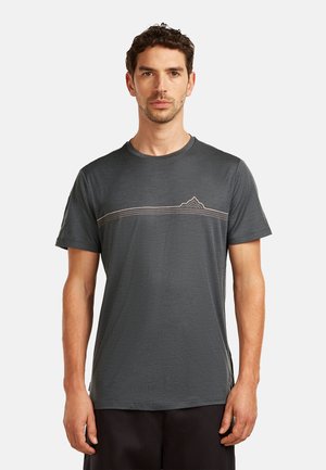 Man staat frontaal en draagt een donkergrijs kort shirt met een dunne horizontale berglijn over de borst.