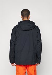 Zwarte waterdichte jas met een capuchon, voorzien van een ontspannen pasvorm en subtiele stikdetails, gecombineerd met feloranje broeken.
