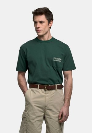 Uomo che indossa una t-shirt verde scuro con tasca frontale e pantaloncini cargo beige, in piedi con una mano in tasca su sfondo bianco.