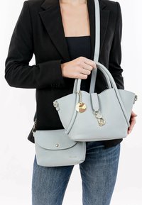 Lichtblauwe leren handtas met textuurafwerking, dubbele handvatten, gouden hardware en een klein extra zakje, gedragen naast een zwarte blazer.