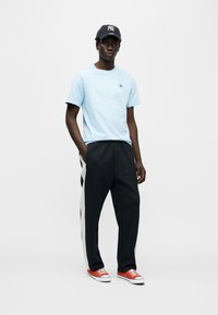 Ανοιχτό μπλε βαμβακερό μπλουζάκι με λογότυπο, μαύρο παντελόνι με λευκές ρίγες στα πλάγια και κόκκινα καμβά παπούτσια. Καπέλο με μαύρες και λευκές ρίγες.