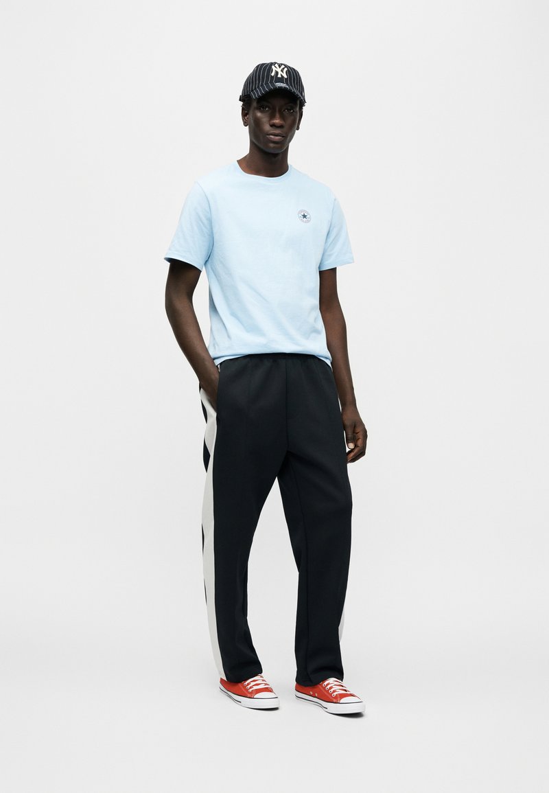 Lyseblå bomulds-T-shirt med et logo, sorte bukser med hvide sidelommer og røde lærreds-sneakers. Sort og hvidstribet kasket.