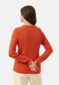 Pull rouge à manches longues en maille douce, avec un col rond et des poignets côtelés, vu de dos avec les mains jointes.