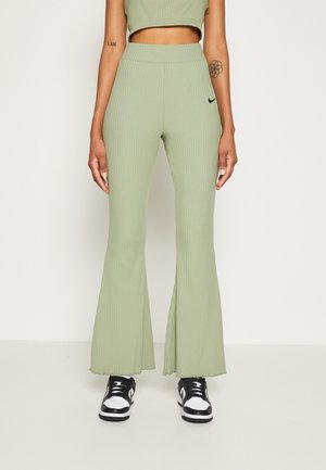 Personne portant un pantalon évasé côtelé vert clair, des baskets noires et blanches, et un crop top avec des tatouages visibles sur les bras, debout sur un sol blanc.