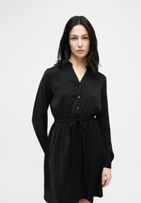 Γυναίκα με σκούρα μαλλιά που φοράει μακρυμάνικο μαύρο φόρεμα με κουμπιά και δεμένη μέση, στέκεται μπροστά σε απλό φωτεινό φόντο.