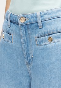 Ανοιχτό μπλε τζιν με ψηλή μέση, μπροστινές τσέπες με χρυσά κουμπιά ως διακοσμητικά και τυπική ραφή κατά μήκος των ραφών.