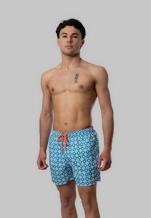Giovane uomo in piedi a torso nudo che indossa pantaloncini da bagno blu a motivi con coulisse arancione, sfondo grigio neutro.