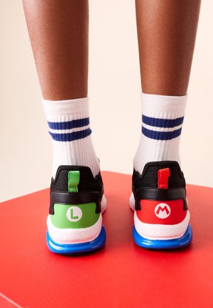 Persoon draagt witte sokken met blauwe strepen en zwarte sneakers met groene "L" en rode "M" logo's op de hielen, staand op een rood oppervlak.