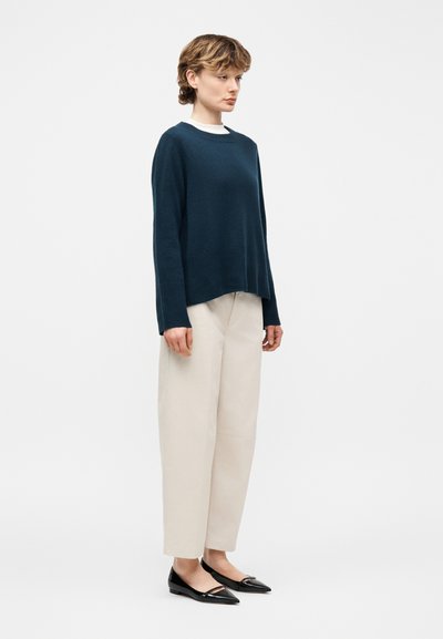 Γυναίκα που στέκεται με μπεζ παντελόνι φαρδύ στο κάτω μέρος, μπλε σκούρο πουλόβερ με μακριά μανίκια πάνω από λευκό τοπ και μαύρες μυτερές μπαλαρίνες, μπροστά από απλή φόντο.
