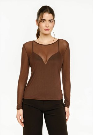 Donna con capelli scuri legati indietro che indossa una maglia marrone trasparente a maniche lunghe sopra una canottiera abbinata e pantaloni neri, in piedi davanti a uno sfondo bianco.