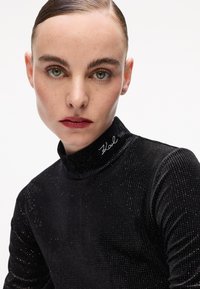 Donna con capelli lisci e indietro che indossa un dolcevita nero glitterato con la scritta "Karl" ricamata sul colletto, che guarda dritto davanti a sé con un'espressione neutra.