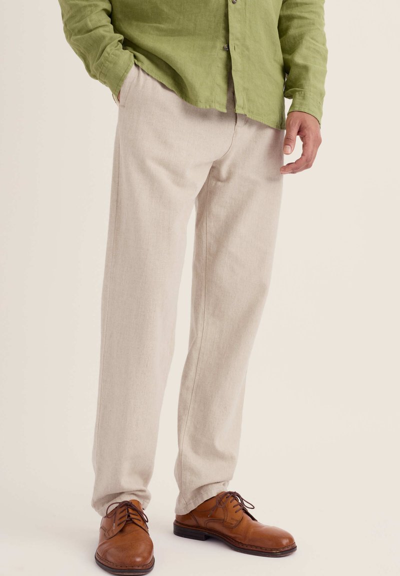 Homme portant un pantalon beige, des chaussures en cuir marron à lacets, et une chemise verte à manches longues, une main dans la poche.