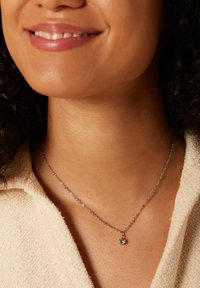 Zilveren ketting met een kleine groene edelsteen hanger, gedragen tegen een lichtgekleurde, gestructureerde top.