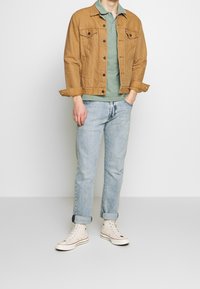 Homme portant une veste en denim beige, un polo vert, un jean bleu clair avec des revers, et des baskets montantes blanches, se tenant contre un fond uni.
