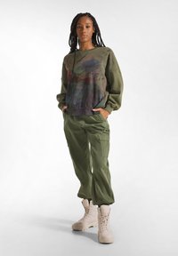 Olivgrüner Sweatshirt mit abstrakten Mustern, kombiniert mit passenden Cargo-Hosen. Helle beige Schnürstiefel runden das Outfit ab.