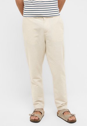 Beige, leichte Hose mit entspannter Passform, hergestellt aus glattem Material und mit Seitentaschen. Kombiniert mit grauen Sandalen mit Schnallenriemen.