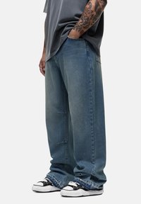 Jeans in denim blu larghi con design a cinque tasche, caratterizzati da una gamba dritta e un leggero sbiadimento. Abbinati a sneakers nere e bianche.