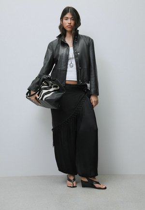 Femeie purtând o jachetă neagră de piele, top alb, pantaloni largi negri cu franjuri, ținând o geantă cu imprimeu de zebră și purtând sandale negre cu platformă.