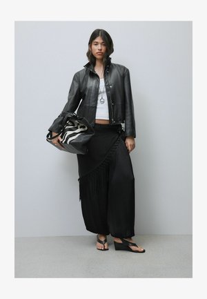 Woman wearing black leather jacket, white top, black wide-leg pants with fringe, holding zebra-print bag, and wearing black wedge sandals.